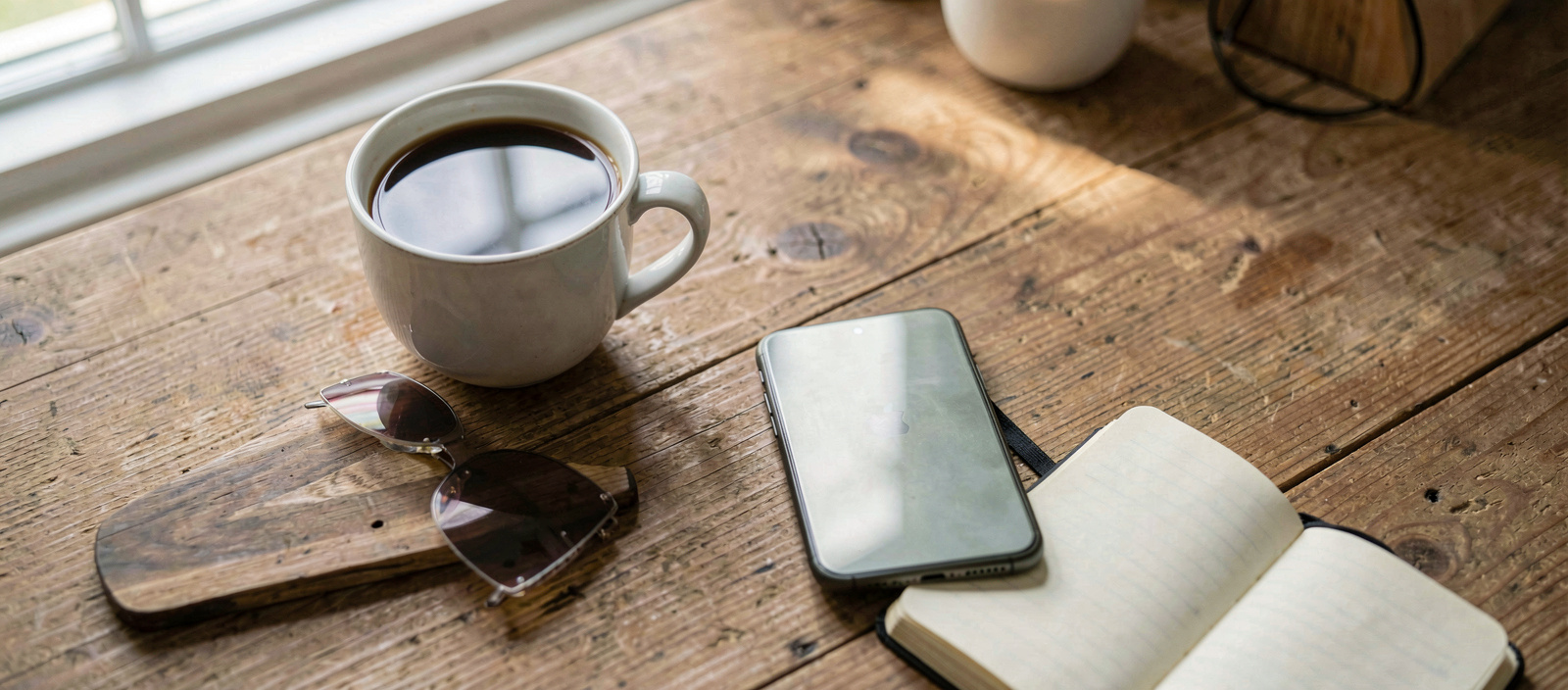 A smartphone lying on a wooden kitchen table next to a cup of coffee and a small notebook, morning light from a window falling across the screen, close overhead framing.
