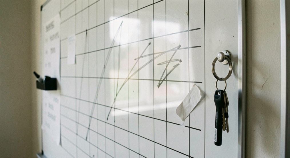 A wall whiteboard covered with a rota grid of half erased lines, a black marker in its tray and a key ring on the nail beside it