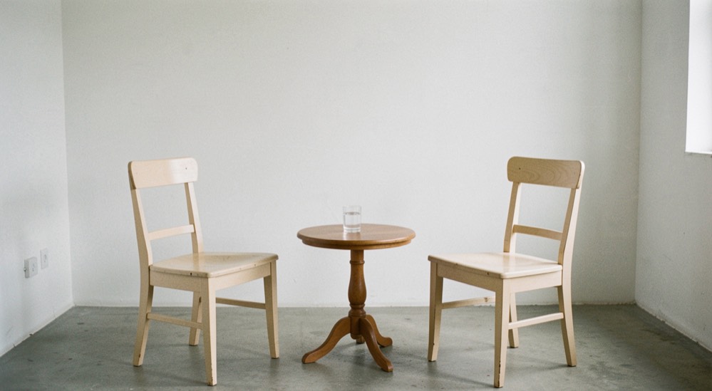 Two pale wooden chairs facing each other in an empty white walled room, a small round table between them with a glass of water