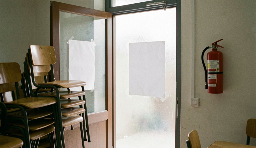A classroom door ajar on stacked chairs, a blank poster taped to the frosted glass and a red extinguisher on the wall
